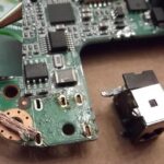 Laptop motherboard with a detached DC charging port removed from the board, requiring solder repair or charging port replacement.